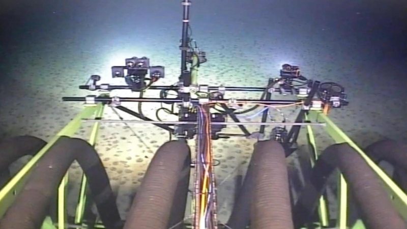 Ein Tiefsee-Bergbau-Roboter sammelt auf dem Meeresboden Manganknollen. Es sind Arme, Kabel und Lampen des Roboters zu sehen, die den Meeresboden voraus beleuchten und durchsuchen.
