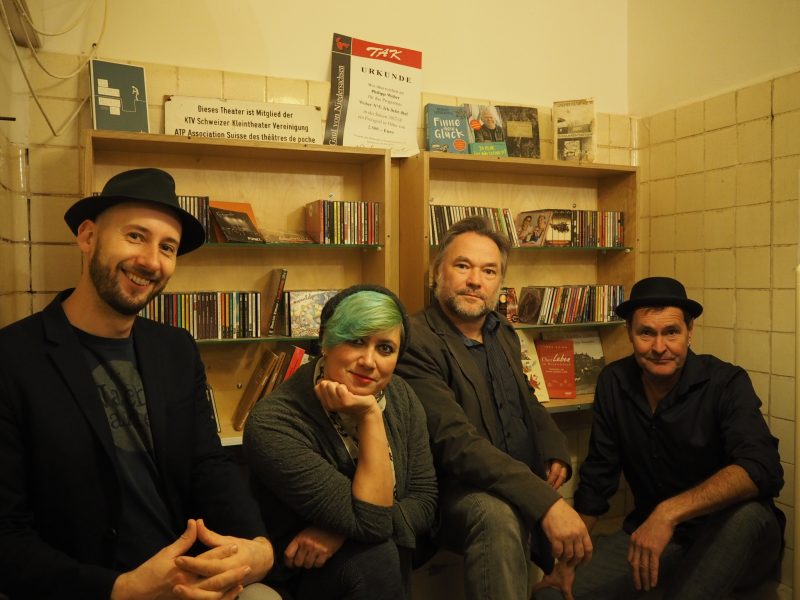 Die Nachtbarden (Tobias Kunze, Johannes Weigel, Kersten Flenter und Ruby S. Zeugs) sitzen in dunkler Kleidung in einem gefliesten Backstagebereich vor einem Regal voller Bücher und CDs und schauen uns mit einem Lachen an.