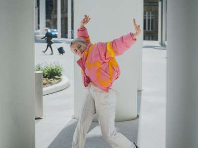 Eine junge blondhaarige Person in weiter, bunter Jacke und heller Hose tanzt zwischen großen Säulen im Freien.