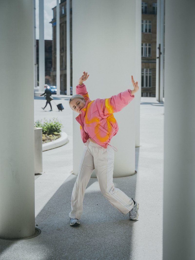 Eine junge blondhaarige Person in weiter, bunter Jacke und heller Hose tanzt zwischen großen Säulen im Freien.