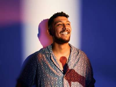 Abdul Kader Chahin (Mann mit dungklen Haaren und Bart) steht in ein sommerliches, kurzärmeliges Hemd mit Muster und eine helle Hose gekleidet im Lichtschein. Er lächelt breit. Auf seiner Brust ist der Schatten einer Hand mit Mikrofon zu sehen.