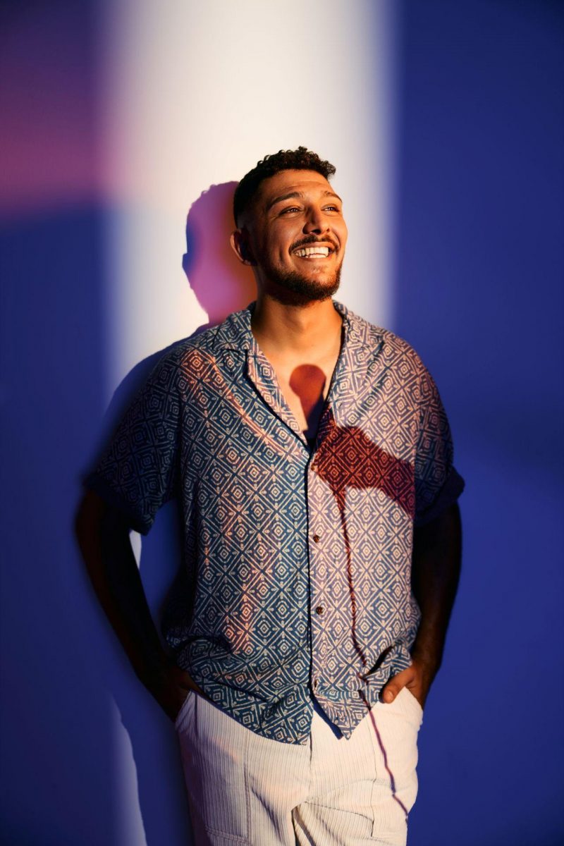 Abdul Kader Chahin (Mann mit dungklen Haaren und Bart) steht in ein sommerliches, kurzärmeliges Hemd mit Muster und eine helle Hose gekleidet im Lichtschein. Er lächelt breit. Auf seiner Brust ist der Schatten einer Hand mit Mikrofon zu sehen.