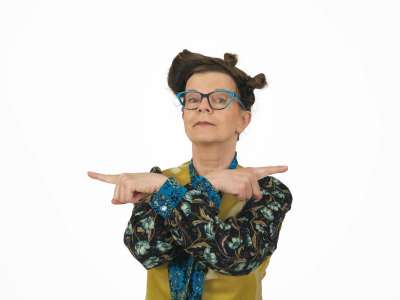 Frieda Braun (Frau mittleren Alters mit bunt gemustererter Kleidung und ausgefallener blauer Brille) zeigt mit beiden Händen in entgegengesetzte Richtungen und schaut dabei leicht verschmitzt in die Kamera.