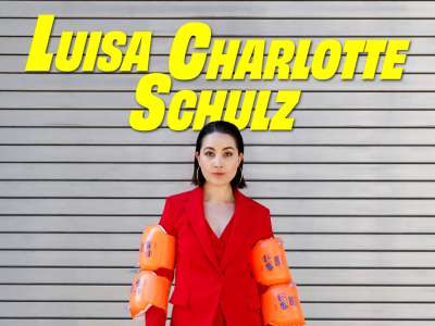 Luisa Charlotte Schulz steht im roten Hosenanzug mit SChwimmflügeln an den Armen vor einem Garagentor. In gelber Schrift ist zu Lesen: Luisa Charlotte Schulz - Stabil instabil.