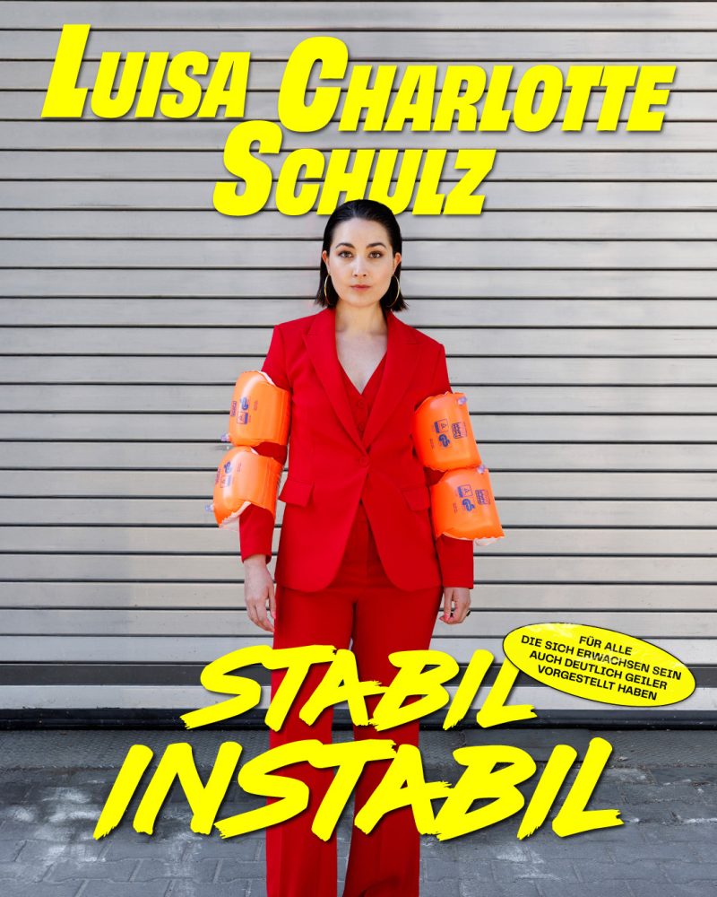 Luisa Charlotte Schulz steht im roten Hosenanzug mit SChwimmflügeln an den Armen vor einem Garagentor. In gelber Schrift ist zu Lesen: Luisa Charlotte Schulz - Stabil instabil.