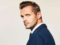 Florian Schroeder schaut über seine Schulter in die Kamera. Er trägt einen blauen Anzug und darunter ein weißes Hemd. Er ist weiß, hat blaue Augen, blondbraune Haare und einen 3-Tage-Bart.