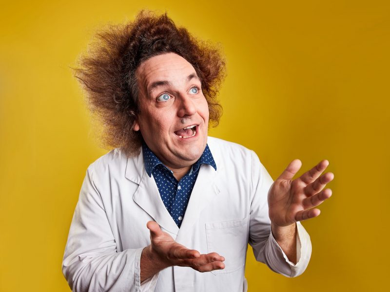 Konrad Stöckel (ein Mann mit zerzausten, zu allen Seiten abstehenden Haaren und einem weißen Kittel an) blickt begeistert nach schräg oben (aus dem Bild heraus) und weist mit seinen Händen in seine Blickrichtung. Hinter ihm ein gelber Hintergrund.