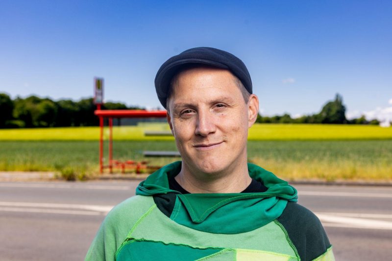 Sebastian 23 (Mann mit grünem Hoodie und schwarzer Schiebermütze) vor einer Landstraße mit roter Bushaltestelle und Feldern