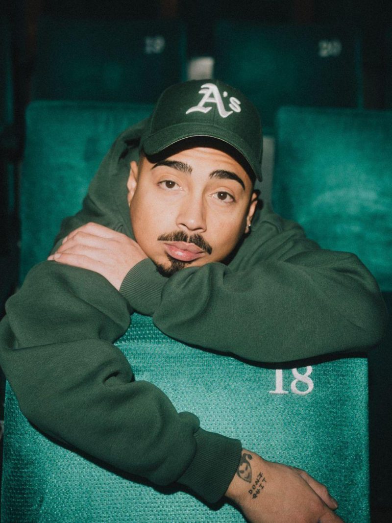 Tony Bauer (mit grünem Hoodie und grüner Baseballcap) lehnt sich nach vorne über einen Theatersitz, legt den Kopf auf seinen linken Arm, während der rechte Arm über die Rückenlehne des vorederen Sitzes baumelt.