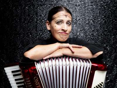 Carmela de Feo (Frau mit dunklen Haaren unter einem Haarnetz, rot geschminkten Lippen und smokey Eyes) lehnt mit verschränkten Armen und einem verschmitzt-herausfordernden Blick auf ihrem roten Akkordeon. Hinter Ihr eine schwarze Wand mit einem im Licht glänzenden Muster.