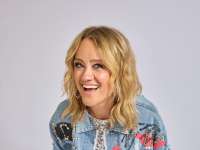 Lisa Feller (Frau mit blonden schulterlangen Haaren, einer blauen Jeansjacke mit farbeigen Details und einem silbernen Paillettenkleid) lacht offen in die Kamera. Der Hintergrund ist weiß/grau.