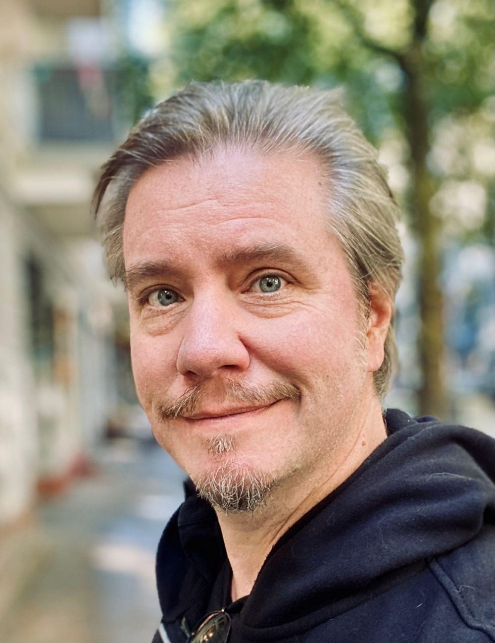 Thilo Gosejohann (Mann mit grauen Haaren, Schnurr- und Kinnbart, blauen Augen und in schwarzem Kapuzenpullover) blickt freundlich in die Kamera. Hinter ihm ist verschwommen eine Straße mit Häusern und Bäumen zu sehen.