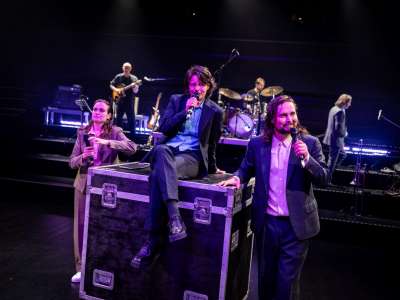 Drei Personen in Anzügen und Hemden sind im Bildvordergrund. Zwei stehen rechts und links eines großen Flightcase (Koffers), die dritte Person sitzt mit überschlagenen Beinen darauf.Alle haben Mikrofone in der Hand. Im Hindergrund ist eine Band zu sehen.