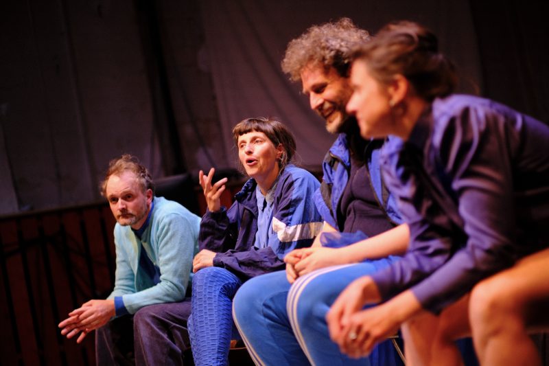 Vier Schauspieler*innen sitzen nebeneinander auf einer Bühne, alle tragen blaue Kleidung, eine Person spricht gestikulierend