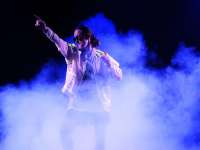 In lila-bläulichem Nebel steht ein*e Performer*in und zeigt mit gestrecktem Arm und Zeigefinger nach oben.