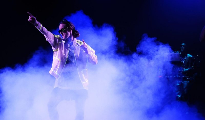 In lila-bläulichem Nebel steht ein*e Performer*in und zeigt mit gestrecktem Arm und Zeigefinger nach oben.